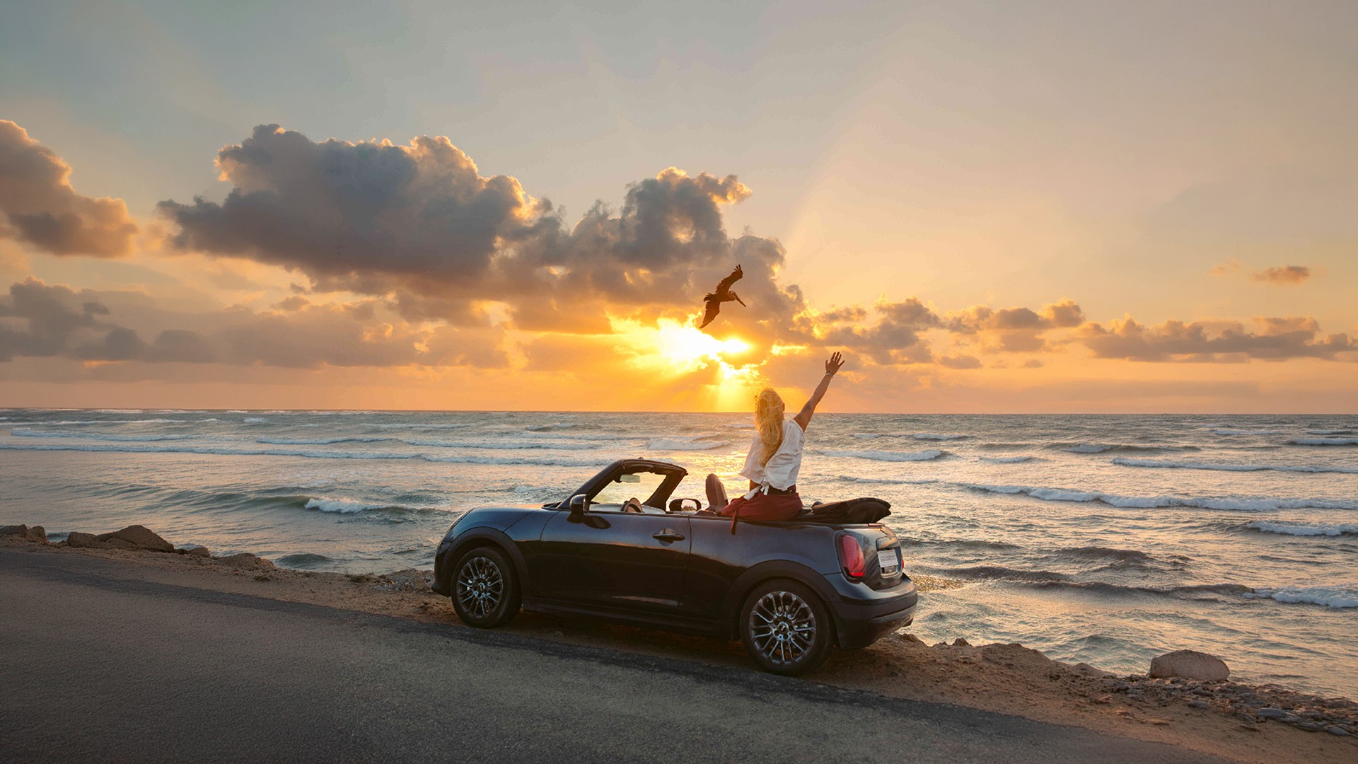 Front left view of the MINI Cooper Convertible in Copper Grey at a beach with two people next to it.